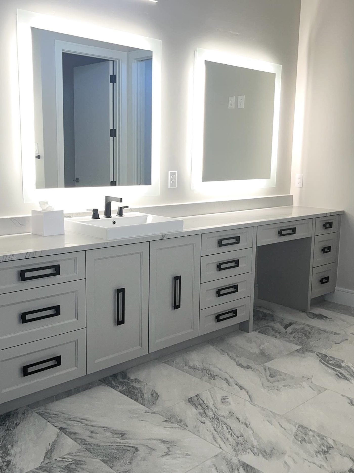 A double vanity with backlit mirrors, gray cabinetry and a marble tile floor