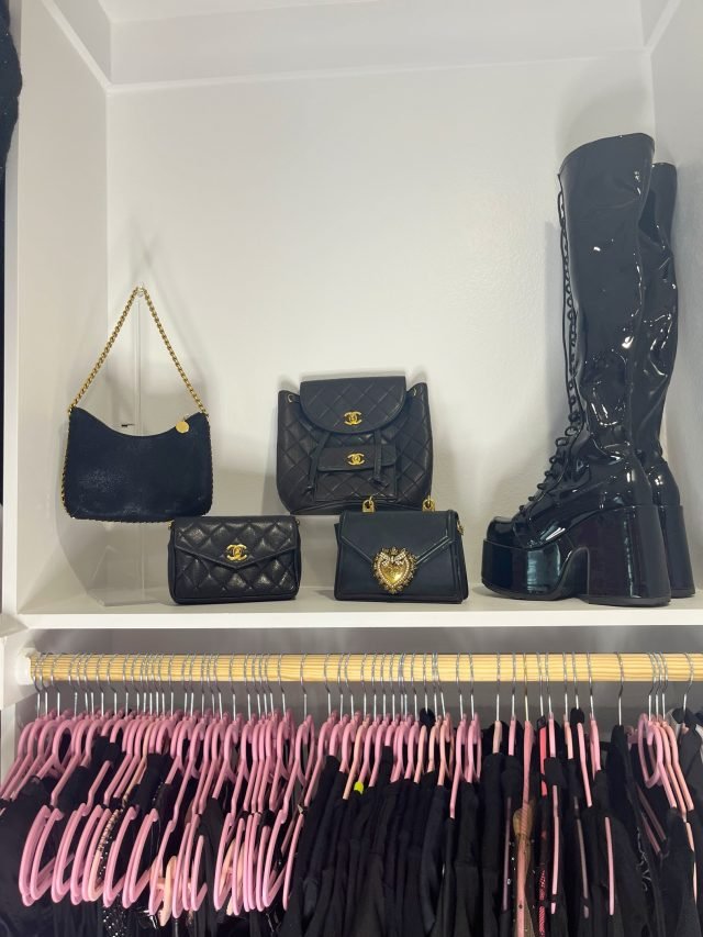 A shelf of black designer handbags and patent boots above a row of hanging pink clothing