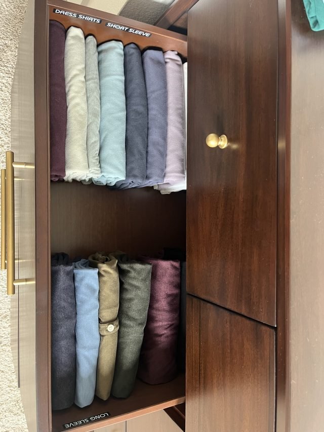 A dark wood closet with T-shirts rolled on edge and jeans rolled below, shelves labeled