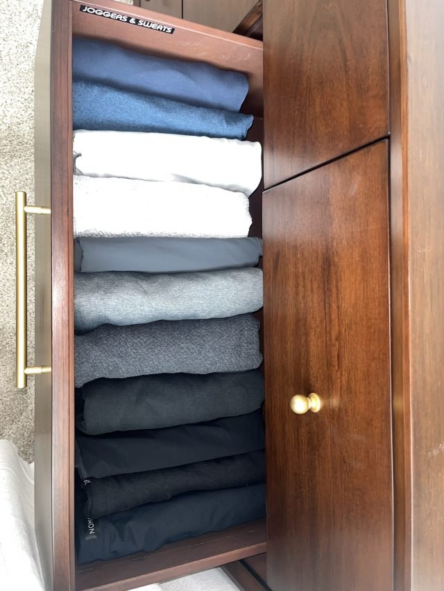 Folded sweaters and white tees stacked by color in a dark wood closet