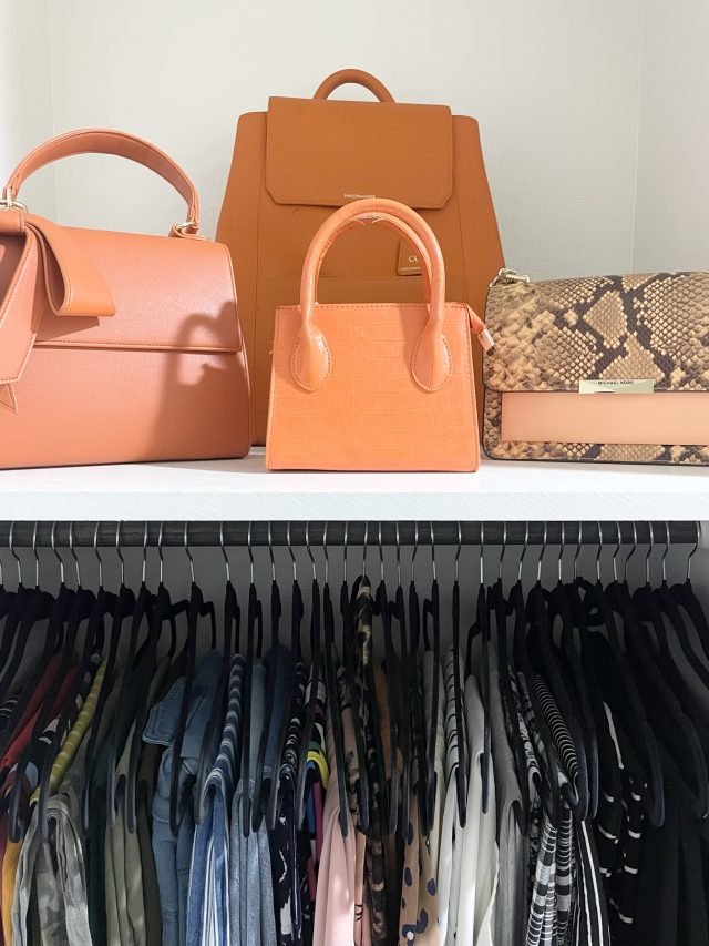 Structured handbags displayed on a shelf above a row of hanging black clothing