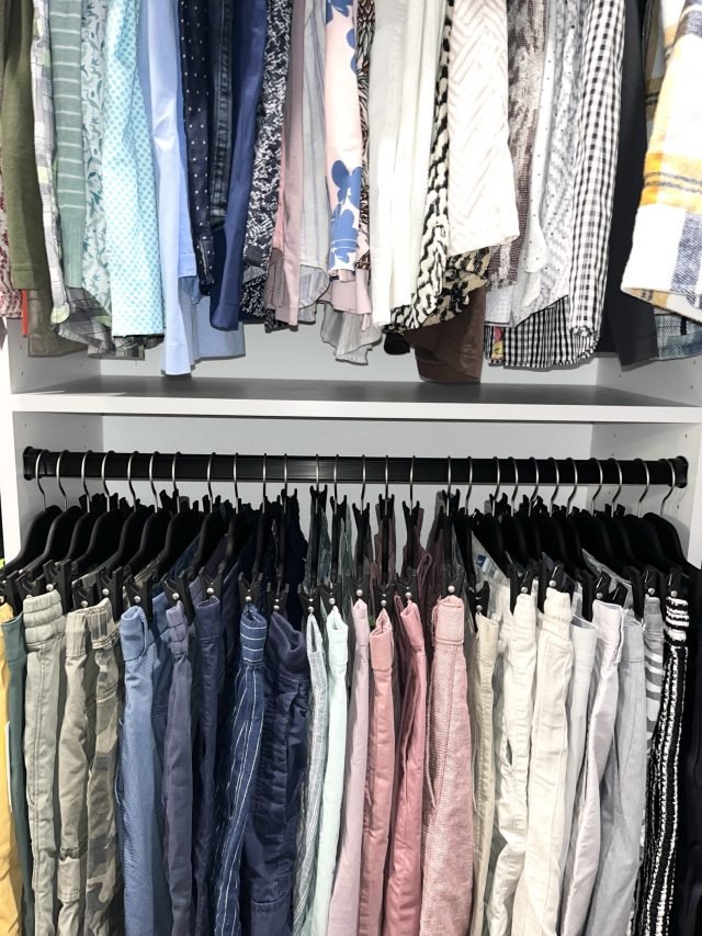 Folded blouses on a shelf above a row of hanging jeans and pants by shade