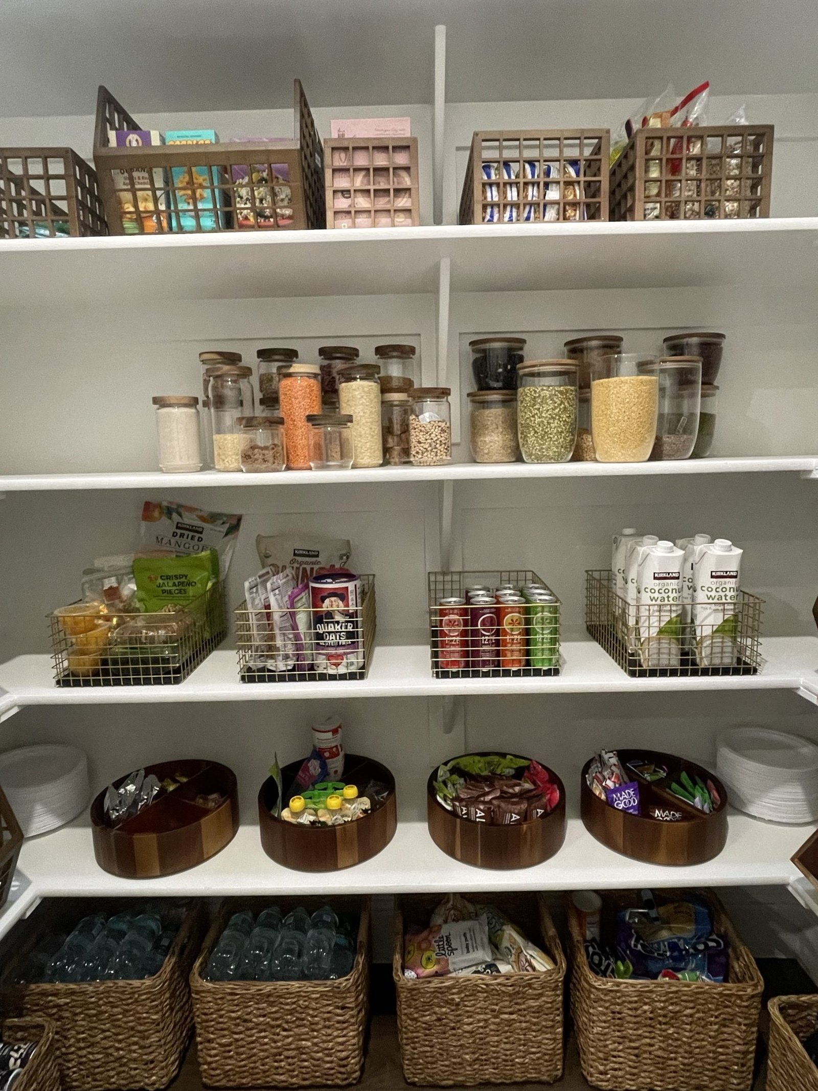 A pantry with dry goods decanted into glass jars, wire baskets of drinks and snacks, and woven baskets on the lower shelf